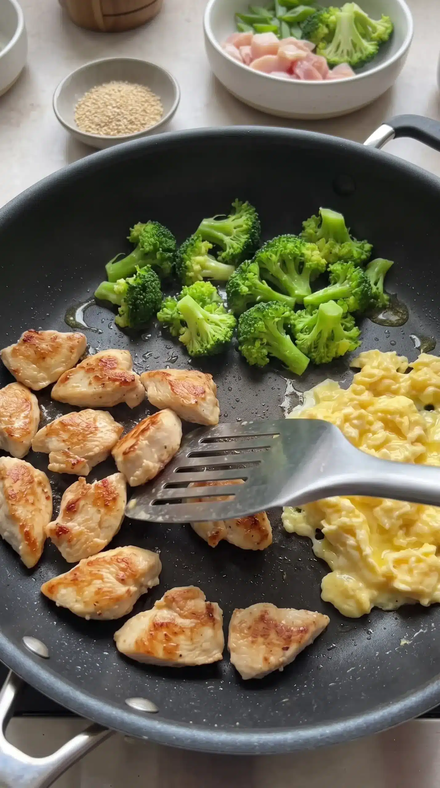Chicken breast slices browning in a skillet with broccoli and eggs being prepared