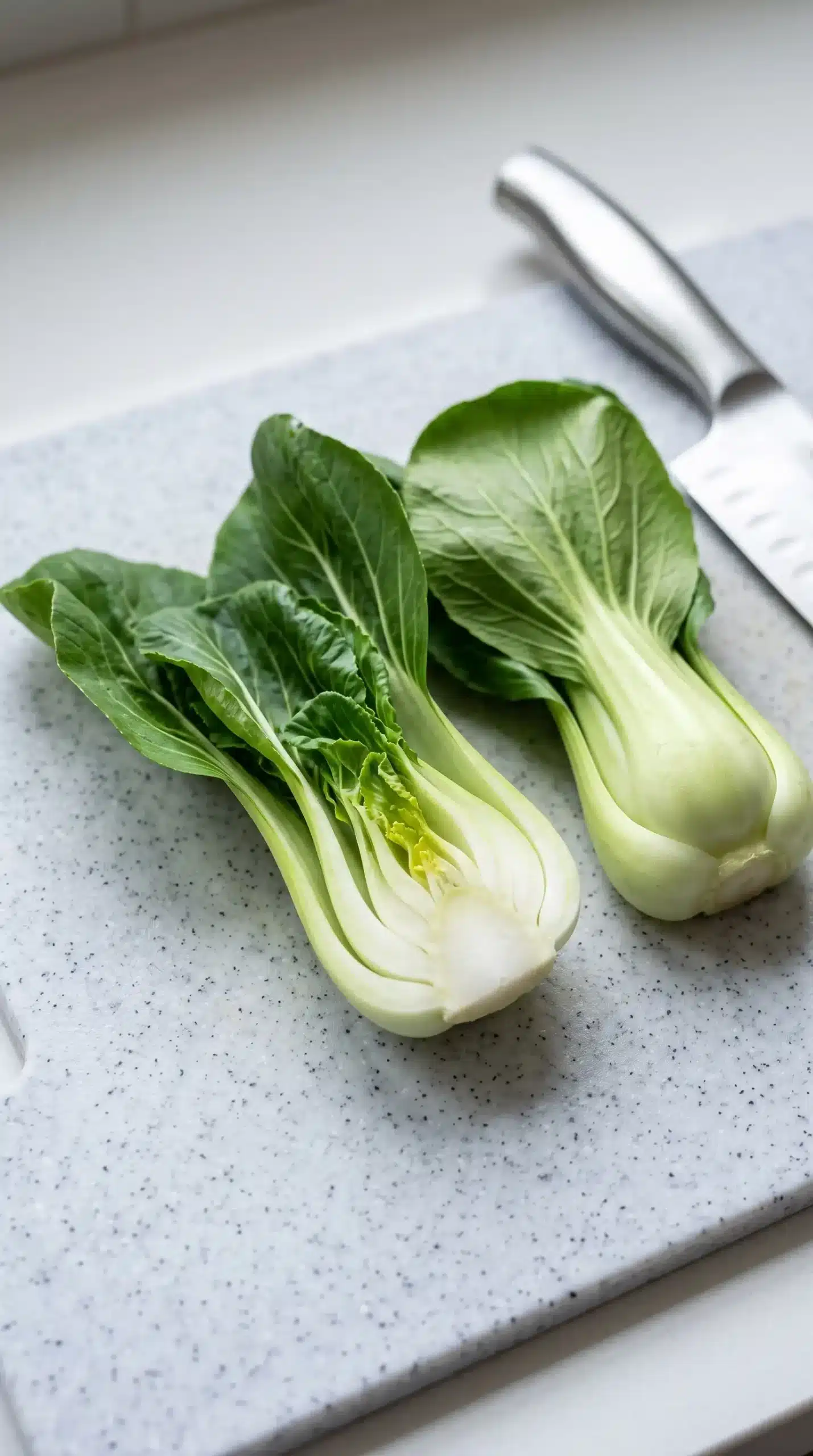 Professional close-up preparation photograph showing fresh bok choy being prepared for bok choy chicken soup: whole baby bok choy and one halved piece revealing the pale crisp stems and green leaves, with a chef's knife and simple cutting surface nearby. Focus on the contrast between stems and leaves and the careful preparation stage before adding to broth. Natural kitchen lighting, realistic food textures, minimal background, no finished dish, no wooden board, no exaggerated arrangement.