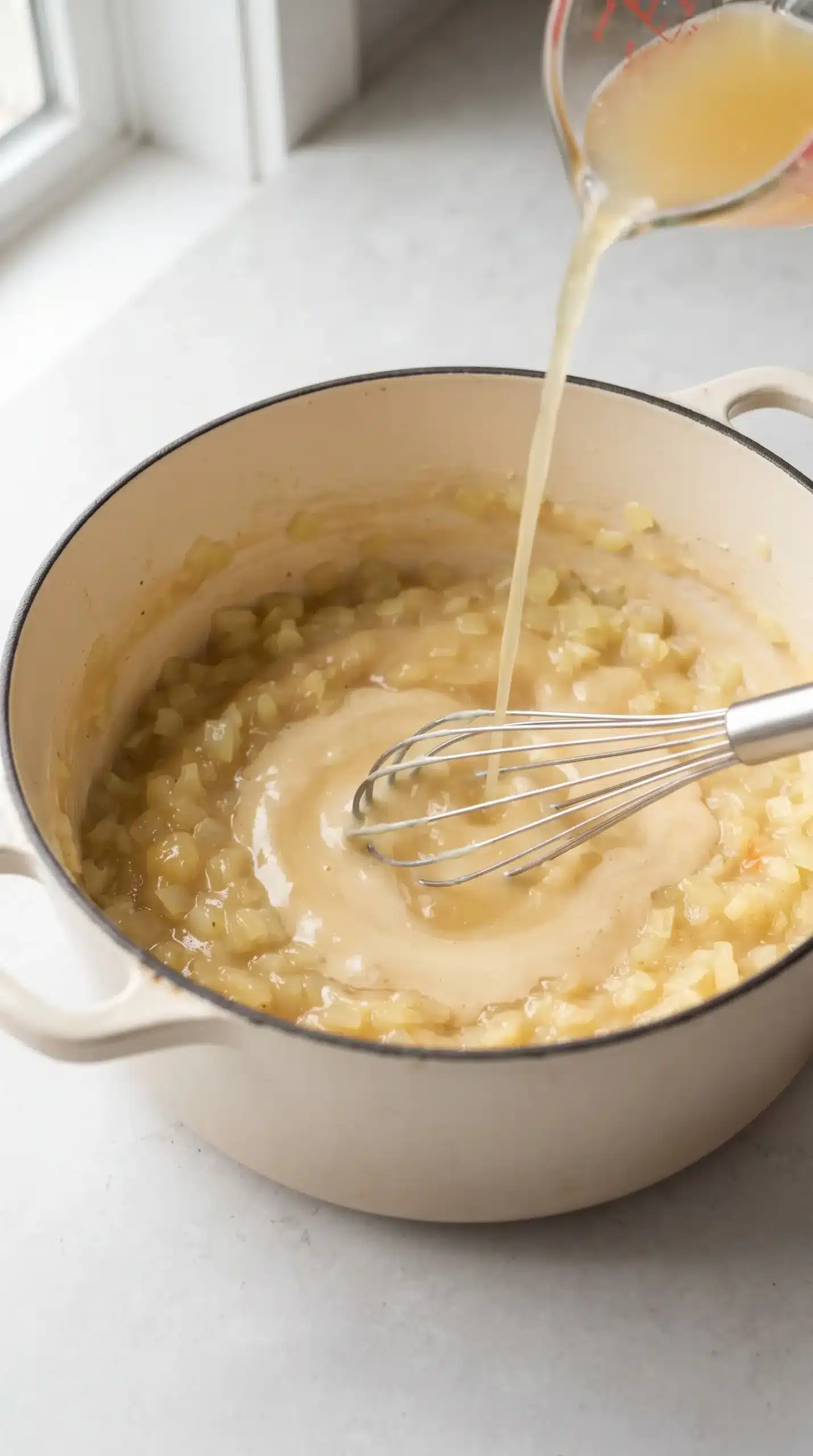Light onion roux being whisked as chicken broth is poured into a cream-colored Dutch oven